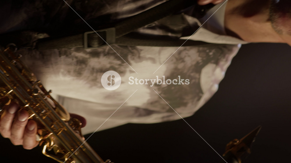 Vertical tilt up low key portrait of middle-aged Caucasian male musician holding saxophone, looking at camera and taking off round glasses, standing on dark background