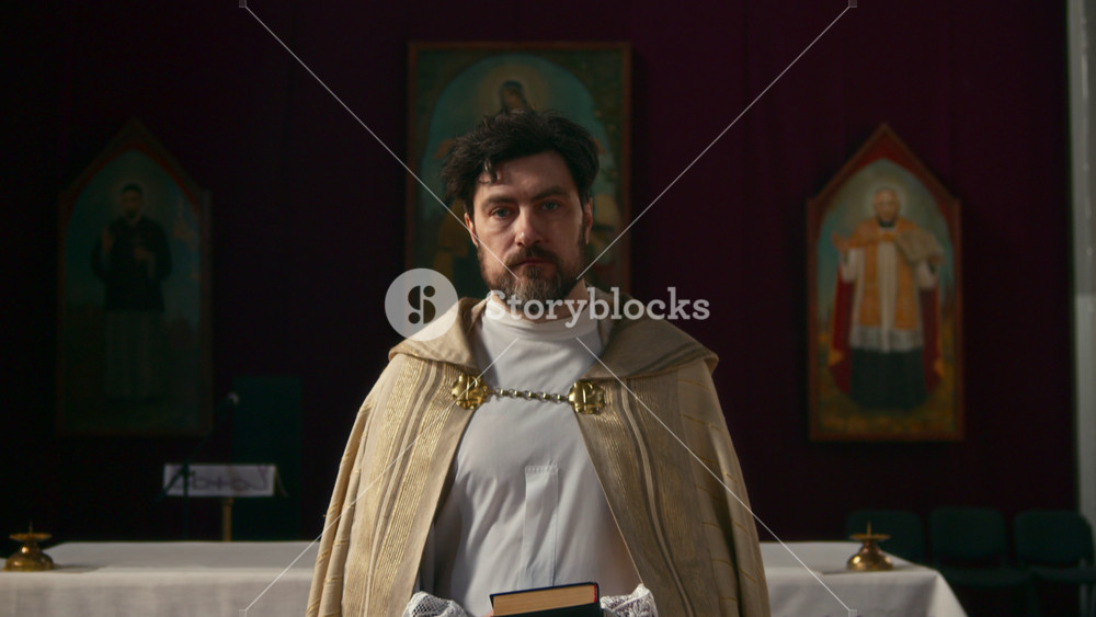 Tilt up portrait of solemn middle-aged Caucasian priest in ceremonial vestment holding Bible looking at camera standing in dim lit church with icons in blurred background