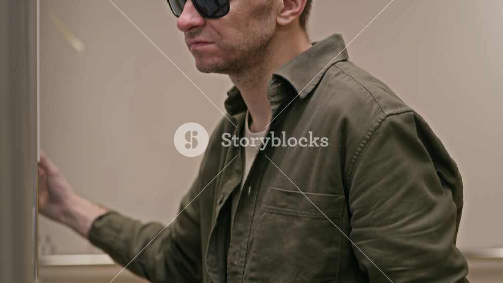Tilt up shot of blind middle-aged Caucasian man in dark glasses holding walking stick standing in front of bookshelf in modern library and choosing braille books, reading tactile text