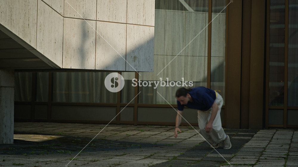 Tilt down shot of skilled young Caucasian guy in streetwear freerunning and performing backflip off concrete wall practicing parkour in urban environment on sunny day