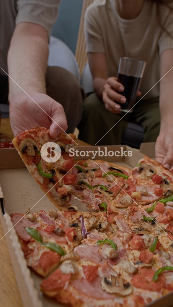 Vertical slowmo of young Caucasian couple taking slices of delicious pizza out of delivery box and clinking glasses with soft drinks, spending time together in modern apartment