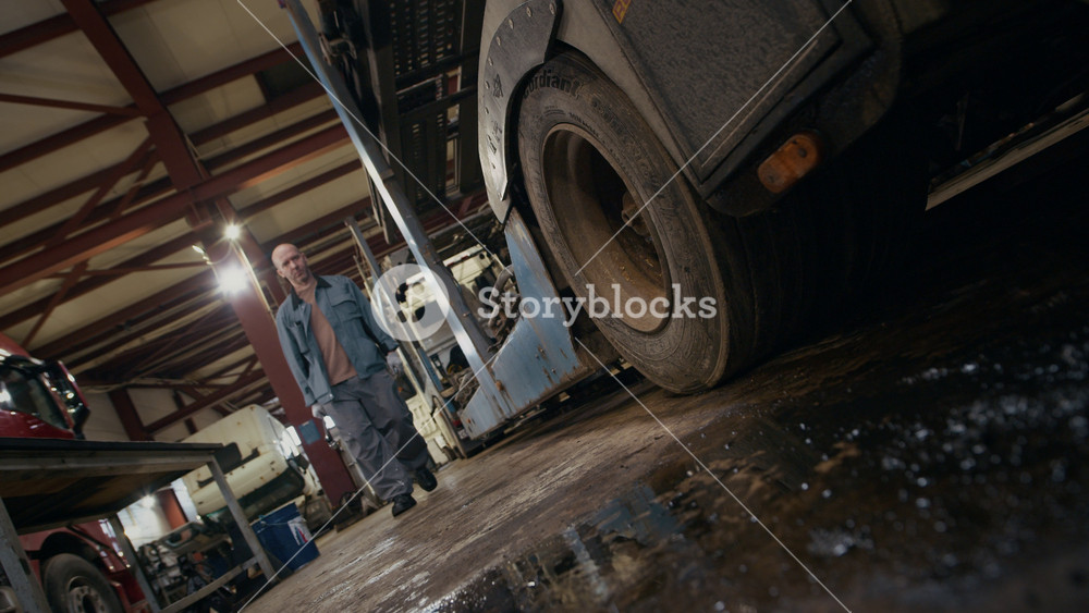 Stab low angle shot of confident middle-aged Caucasian male mechanic holding special car tool approaching truck in auto repair shop and examining its tires