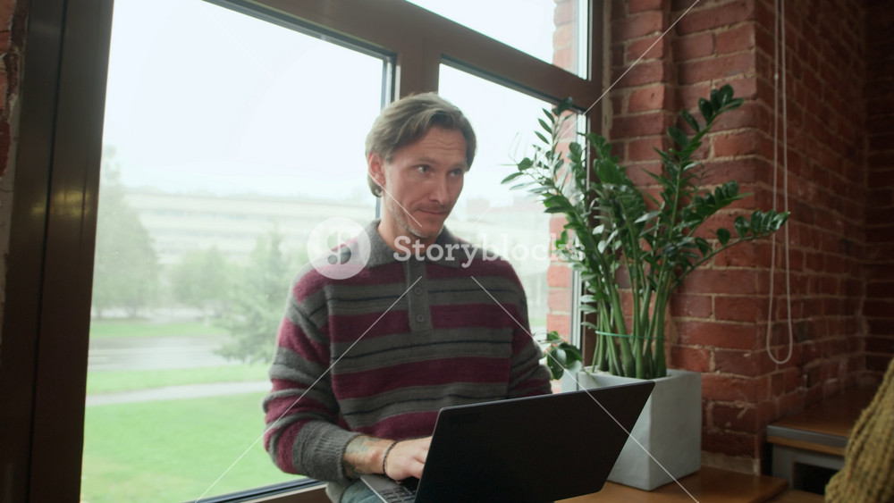 Handheld shot of middle-aged Caucasian man in casualwear greeting his male colleague working on laptop by window while going upstairs in modern office building