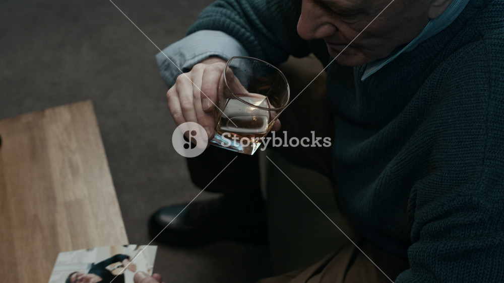 High angle low key shot of sad Caucasian elderly man looking at photo of woman while sipping whiskey, sitting at home and feeling nostalgic about past trying to overcome stress with alcohol