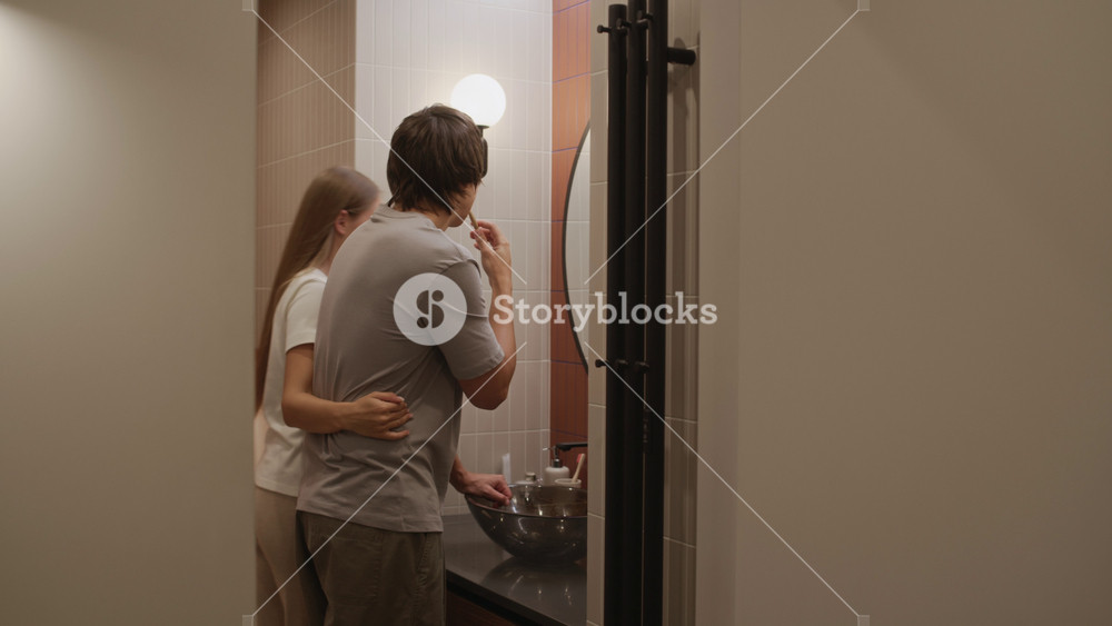 Young beautiful Caucasian woman entering modern bathroom to pick up face cream while her boyfriend brushing his teeth at sink in front of mirror, sharing casual morning moment together