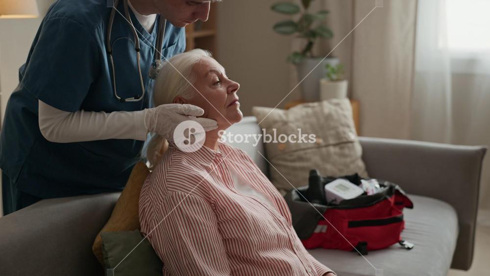 Young Caucasian male nurse checking lymph nodes of senior Caucasian woman during home medical visit. Perfect for promoting geriatric care services and senior healthcare initiatives