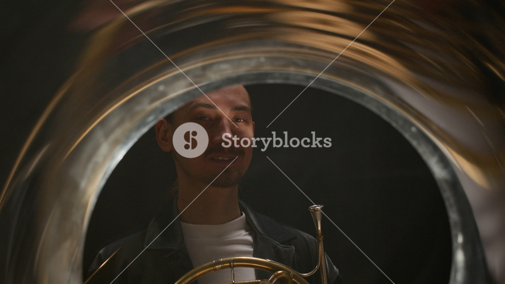 Tilt up portrait of young Caucasian male musician looking at camera with light smile holding French horn, standing on black background being illuminated by moving spotlight