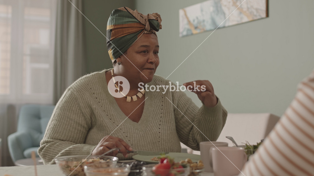Medium shot of beautiful aged African-American woman wearing casual clothes sitting at dining table and having meal with her unrecognizable relatives sitting in front of her