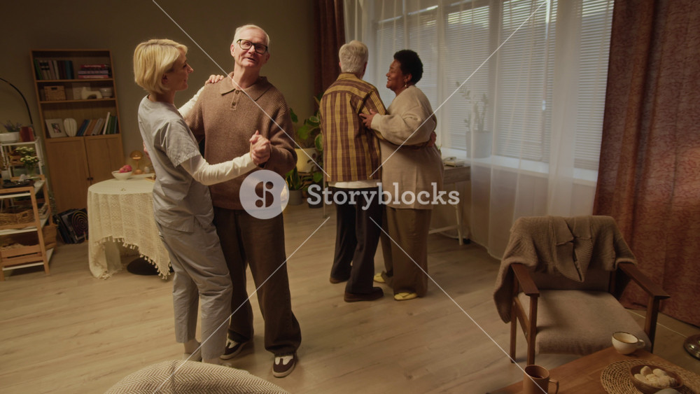 Full slowmo shot of group of diverse elderly residents and female Caucasian caregiver enjoying dancing in pairs in cozy nursing home lounge, sharing moment of connection, movement, and joy