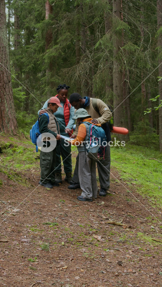 Vertical full shot of African American family of four pausing in forest to examine route map, planning their next steps on hiking adventure together