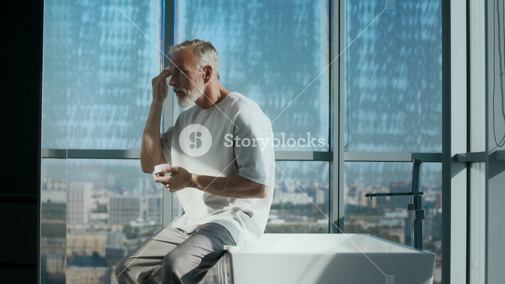 Medium shot of good-looking gray-haired Caucasian adult man dressed in white T-shirt and gray pants sitting on elegant bathtub while applying skincare cream to his face