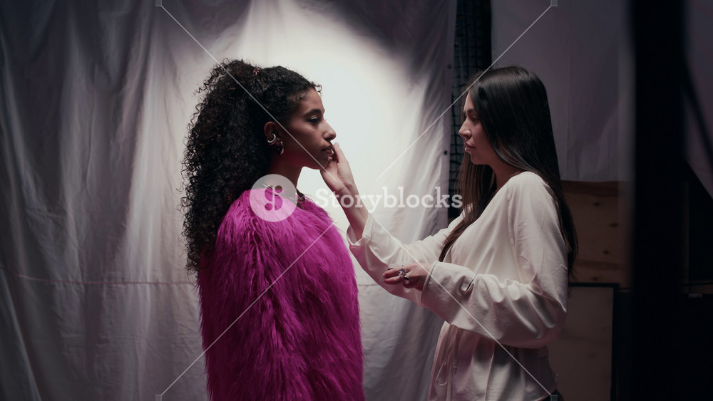 Sideview medium shot of female Caucasian makeup artist applying powder on young beautiful biracial girl working as fashion model wearing pink faux fur coat preparing for studio shooting