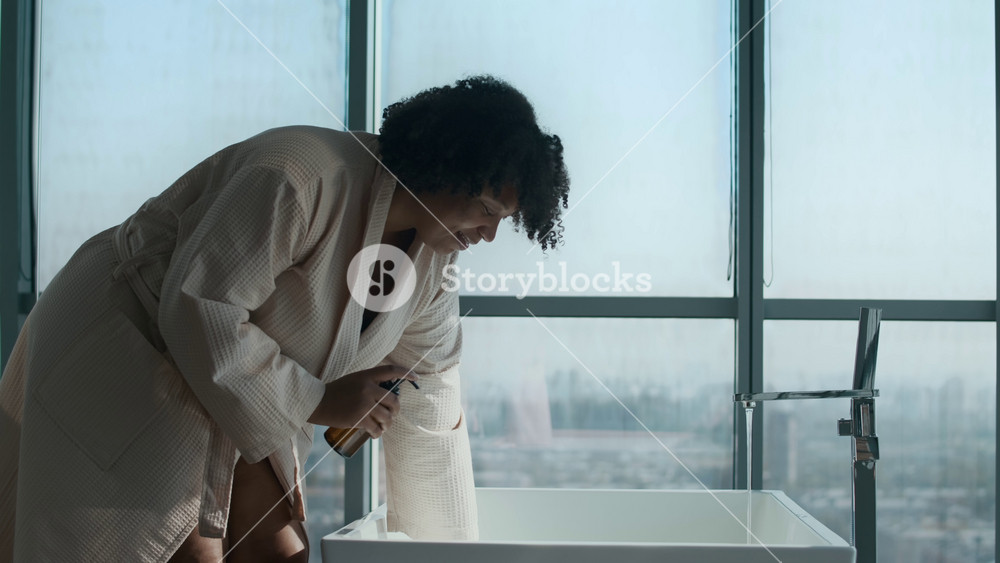 Side view medium shot of young Biracial woman in cozy bathrobe adding some bath oil into filled freestanding bathtub set up on high-rise with stunning city view in modern, luxurious apartment
