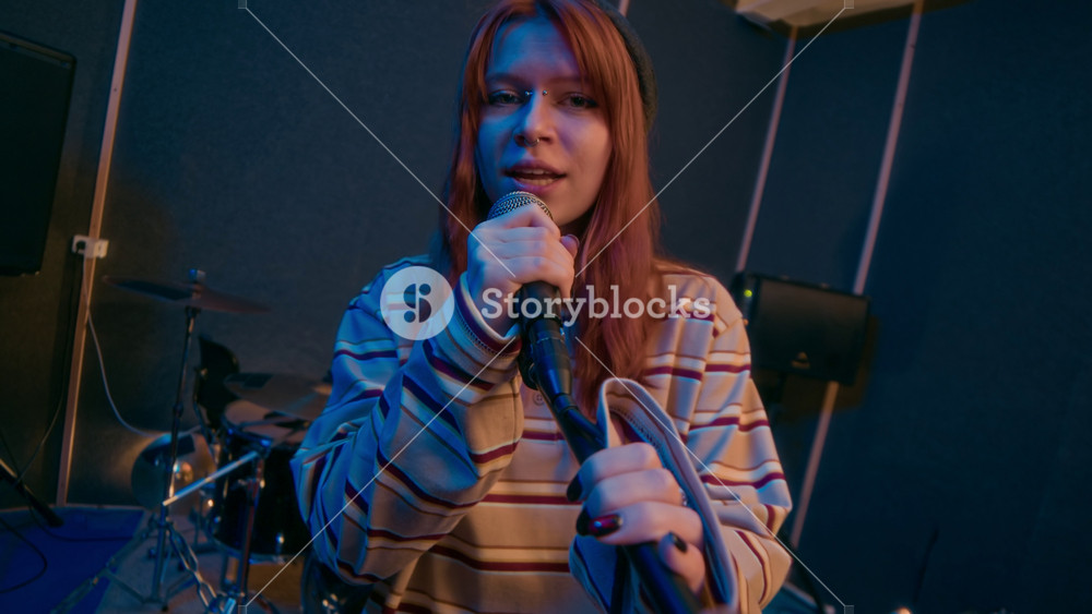 Tracking portrait shot of redheaded punk girl in striped sweater singing rock song into microphone and looking at camera in music studio