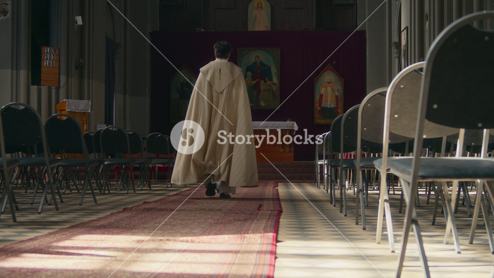 Rear view shot of priest wearing golden festive cope walking along Catholic church among rows of pews heading to altar with icons