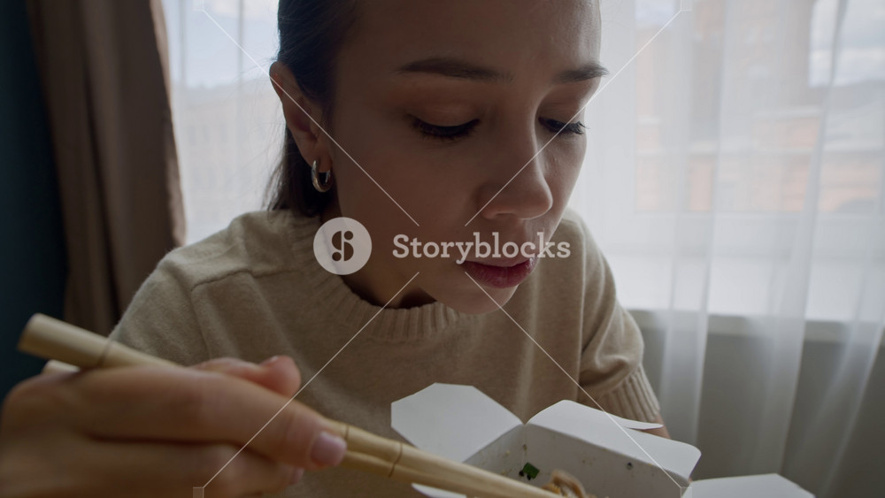 Handheld close up shot of hungry young Caucasian couple chatting while eating wok noodles with chopsticks from cardboard delivery box, having takeout food at home together