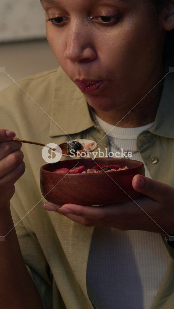 Vertical slowmo tilt up shot of young African American woman eating yogurt bowl with granola and berries, enjoying healthy breakfast rich in fiber and protein