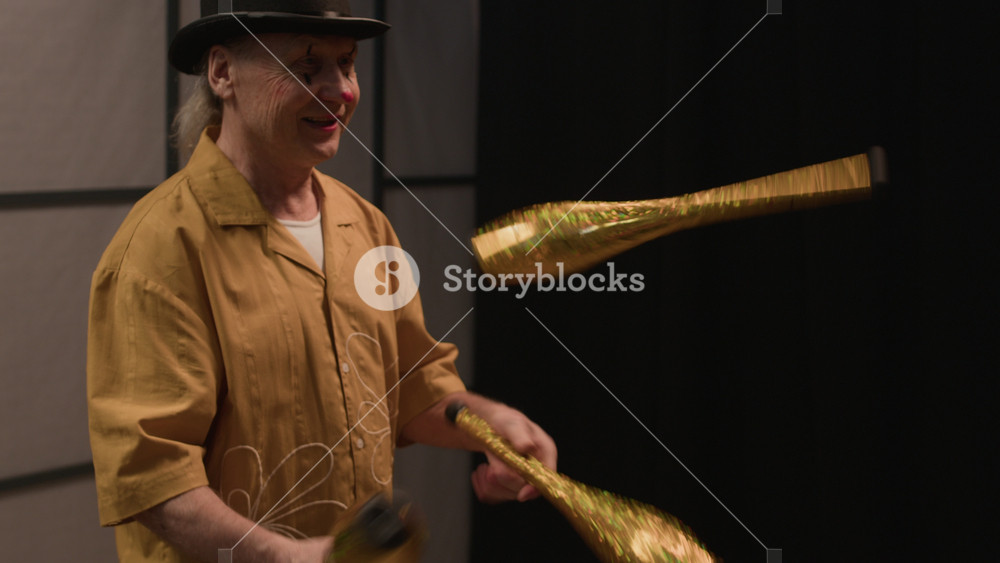 Medium shot of cheerful senior Caucasian juggler in bright yellow costume and black concert hat performing on stage in front of jury during talent show