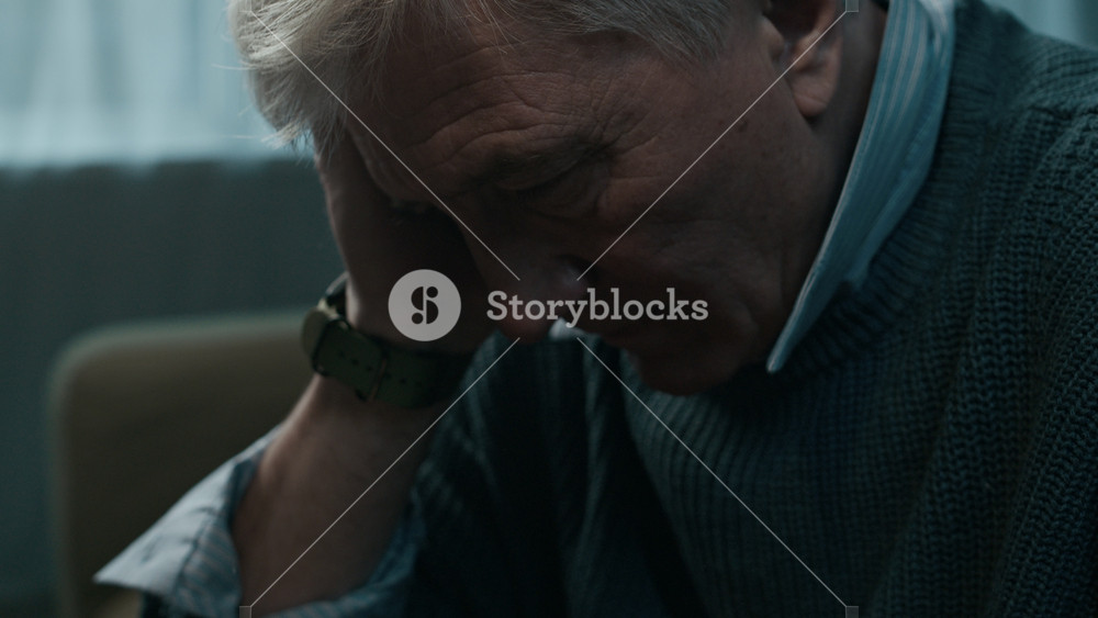 Low key closeup shot of gray-haired senior Caucasian man in despair holding glass of whiskey, rubbing his hair and face trying to overcome crisis and painful emotions with alcohol