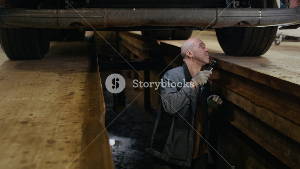 Medium slowmo shot of concentrated middle-aged Caucasian male mechanic examining bottom parts of truck using flashlight, working in auto repair shop