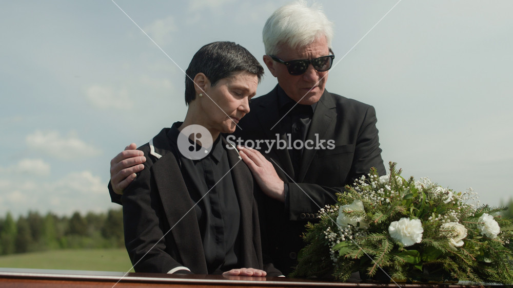 Medium shot of mature Caucasian widow in mourning looking at coffin at outdoor funeral ceremony and being supported by senior man in black suit sorrowing together over death of loved one