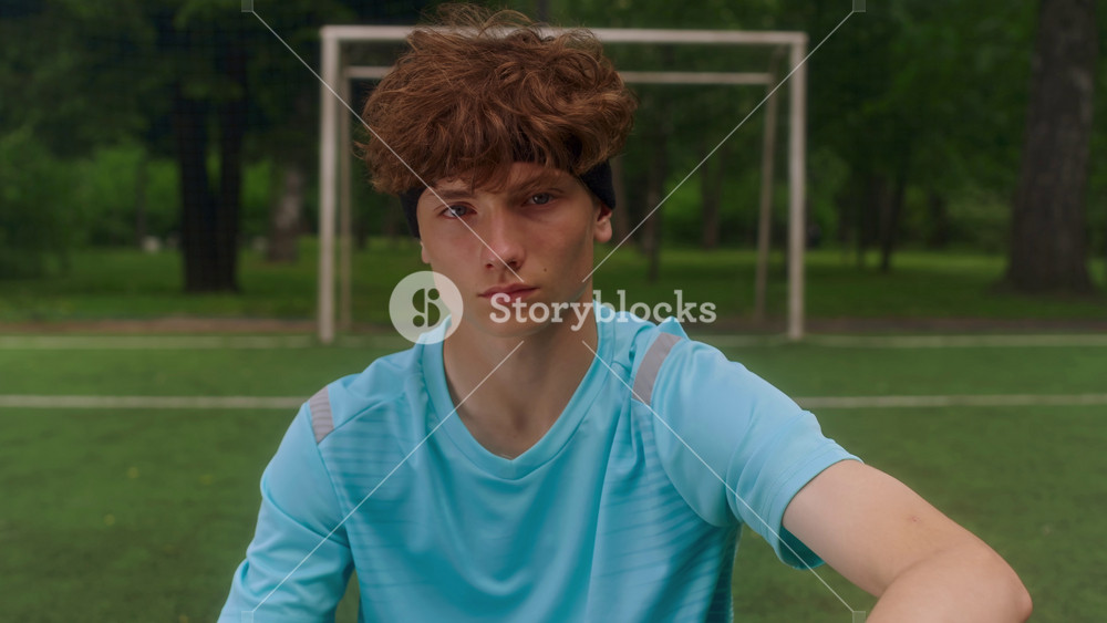 Portrait of Caucasian teen soccer player sitting on grassy field beside ball, looking confidently at camera, wearing soccer gear