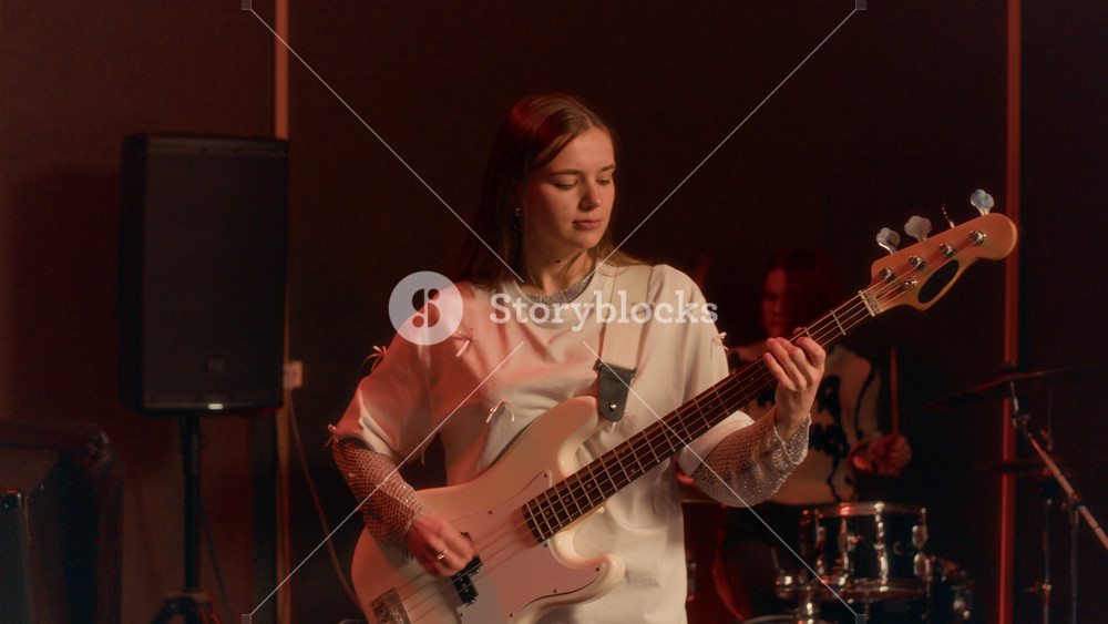Tilt-up shot of young girl in white shirt intently playing white electric bass guitar in dimly lit rehearsal studio, accompanied by female drummer