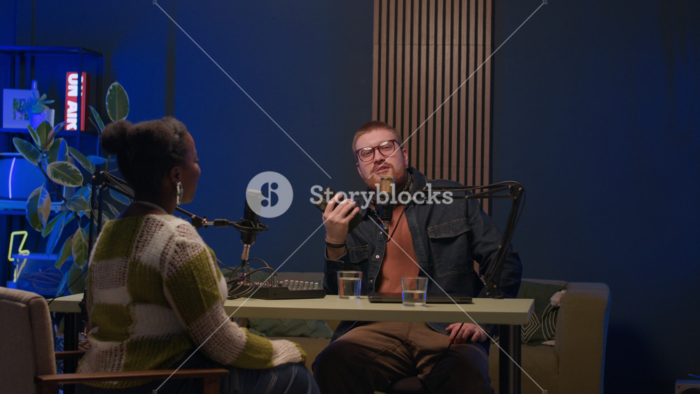 Young Caucasian male podcast host and young Black female guest speaking in mic sitting at table in modern recording studio with neon on air sign shining in dark, then main light turning on