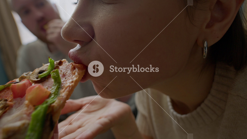 Close up slowmo of young Caucasian woman taking yummy slice out of cardboard pizza box and enjoying fast food at home with her boyfriend eating nacho chips in background