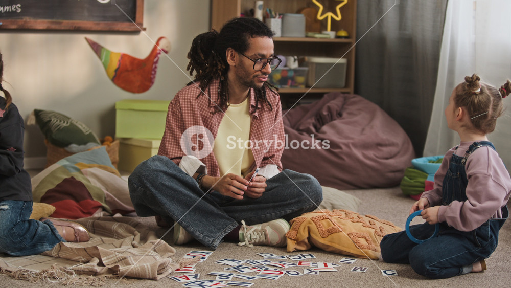 Ground level full shot of motivated young biracial male kindergarten worker teaching alphabet to Caucasian girls using flashcards, sitting on floor in cozy classroom and giving high five