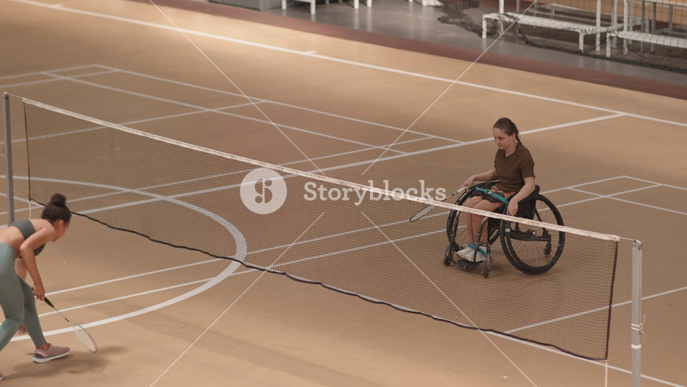 High angle wide shot of Caucasian teenage girl in wheelchair playing badminton with unrecognizable athlete in indoor court