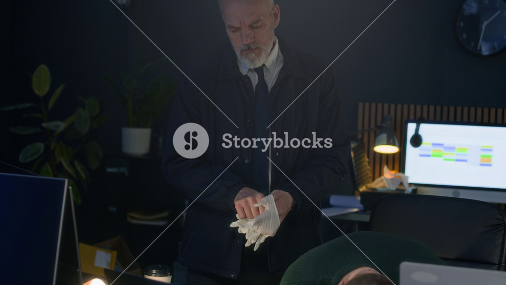 Handheld shot of mature Caucasian male detective in black putting on sterile gloves working at murder scene with unrecognizable colleague, examining male victim lying on open space office desk
