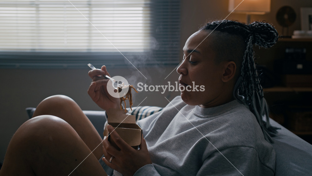 Tilt up of Biracial woman wearing electronic ankle monitor sitting on couch and eating noodles from delivery box while spending time at home