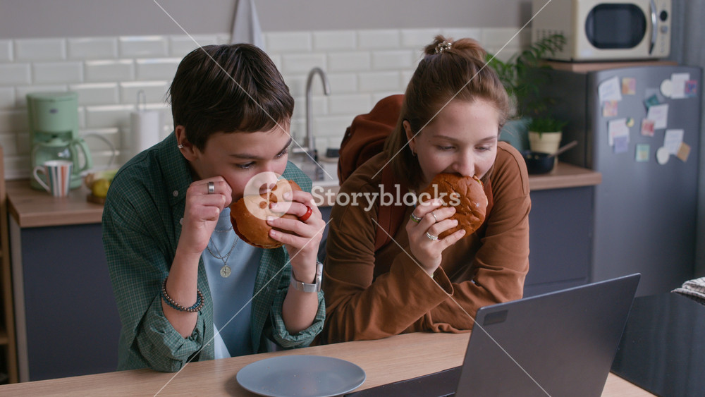 Medium eye level shot of Caucasian teenage boy and girl eating burgers while sitting at kitchen counter with laptop in bright home interior