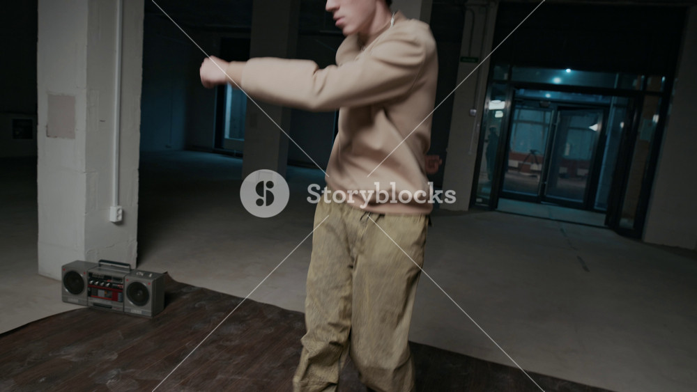Handheld shot of talented Caucasian gen Z b-boy in streetwear performing top rock moves, improvising while breaking alone in indoor dance hall to music on boombox