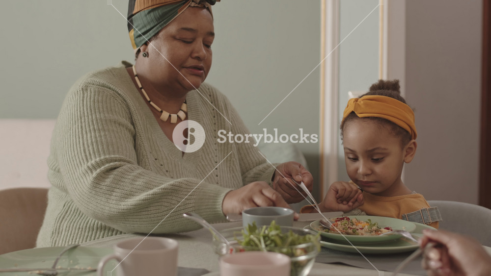 Medium shot of beautiful old African-American woman wearing casual clothes sitting at dining table and trying to feed her little granddaughter who refusing to eat