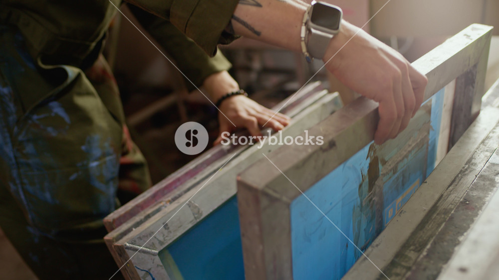 Tilt down shot of Caucasian male worker putting his smartphone in pocket of his coveralls and starting browsing stack of screen printing frames in creative workshop, preparing for branding job
