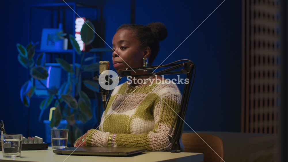 Young Black female podcast guest answering question of young Caucasian male host listening her carefully, both speaking in mic in professional recording studio with black backlight and neon signs