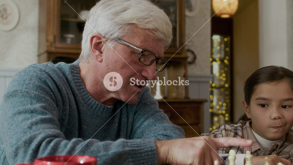 Tilt up of intelligent Caucasian grandfather wearing soft gray sweater and glasses teaching his two grandchildren how to play chess game at wooden table in cozy countryside house