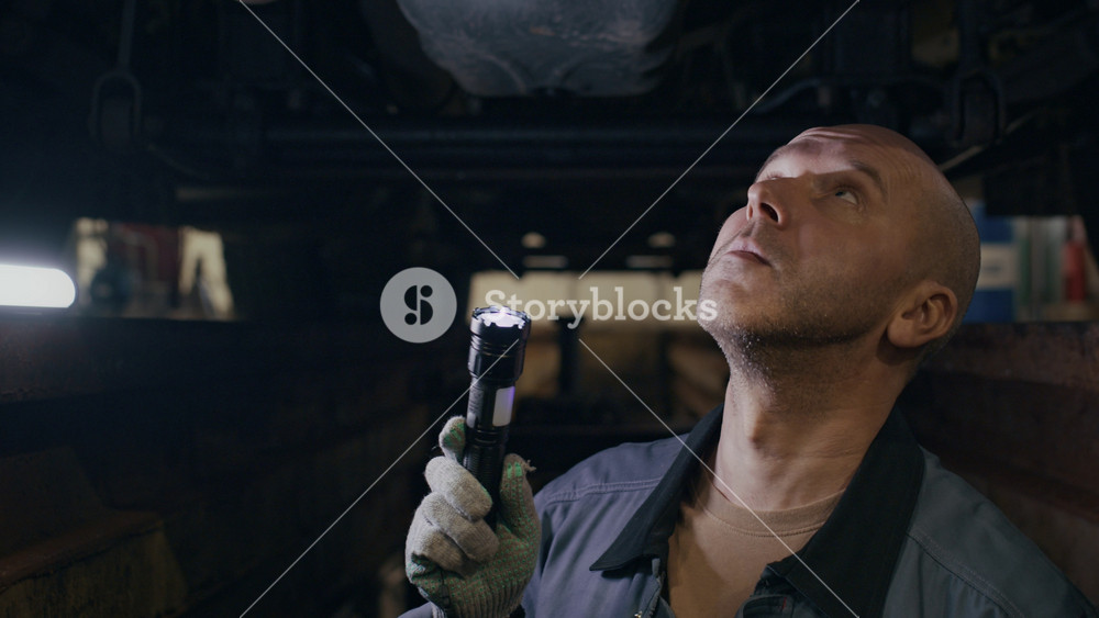 Zoom in shot of concentrated middle-aged Caucasian repairman in jacket and gloves looking up checking bottom parts of truck using flashlight