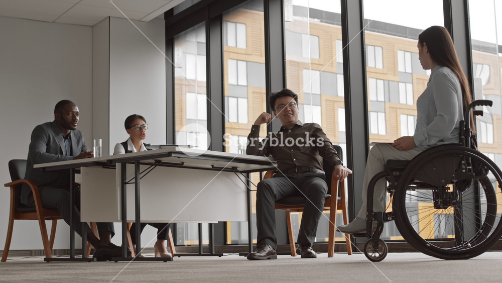 Wide shot of beautiful female businesswoman sitting in wheelchair in modern office and talking to three multiethnic HR managers during her job interview