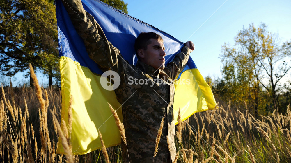 Military man in uniform stands with raised over head flag of Ukraine at countryside. Male ukrainian army soldier with lifted national banner