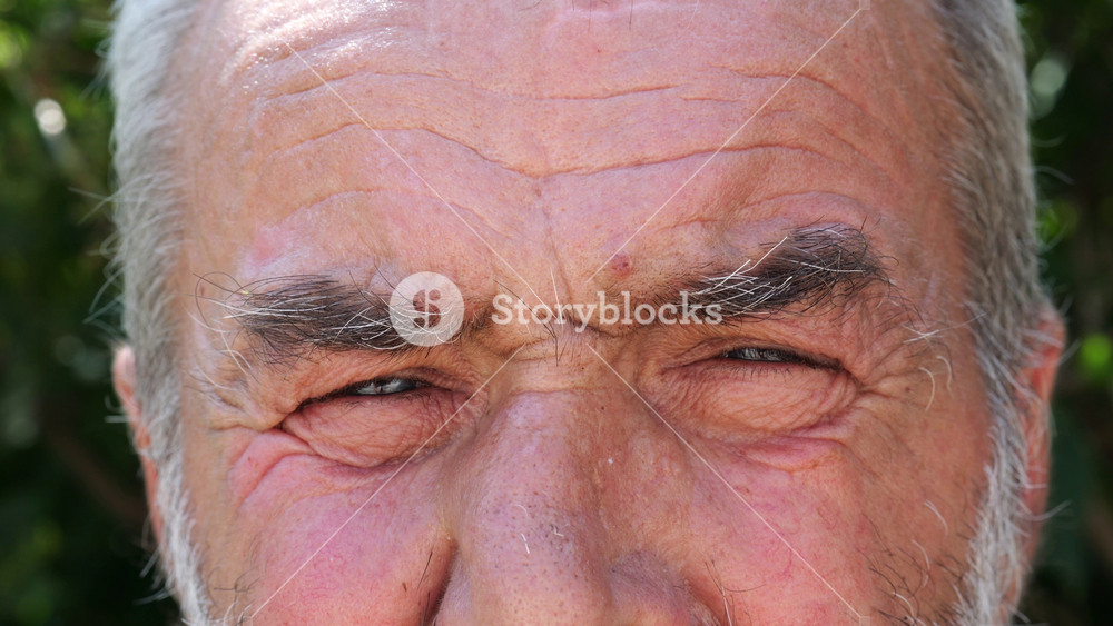Blue eyes of elderly male blinking and looking into camera outdoor. Wrinkled face of senior human with a sad sight. Upset facial expression