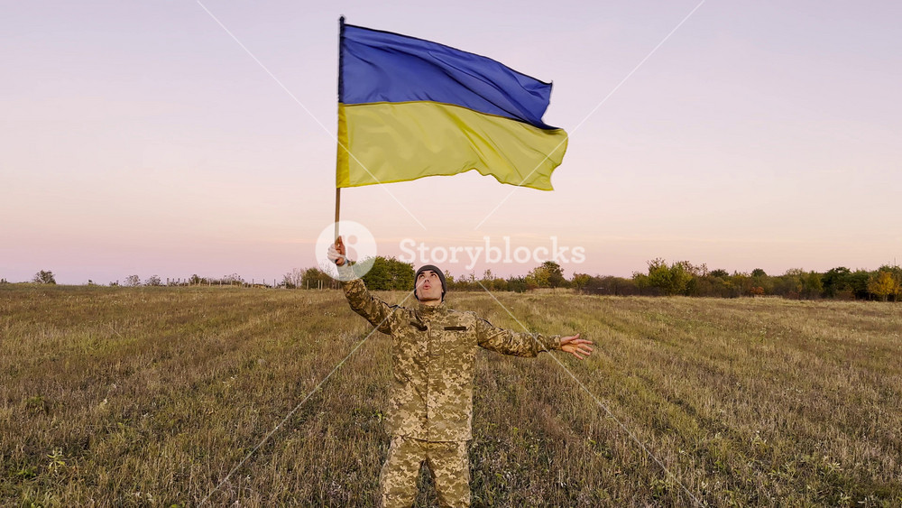 Male military lifted flag of Ukraine in honor of victory against russian aggression at dusk. Young soldier of ukrainian army waving blue