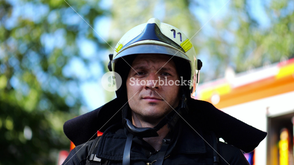 Portrait of young confident fireguard in protective uniform against background of a fire truck. Male firefighter in full equipment looking into camera with positive emotions near big red car. Close up