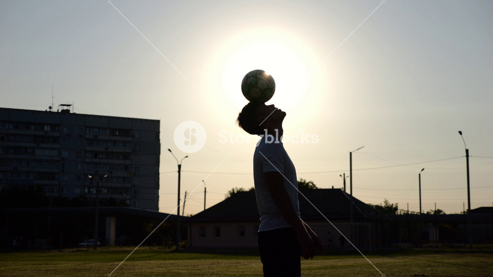 Silhouette of sportsman practicing tricks with ball on head at field on sunset. Professional male footballer in uniform training at meadow with sunlight at background. Freestyle football concept