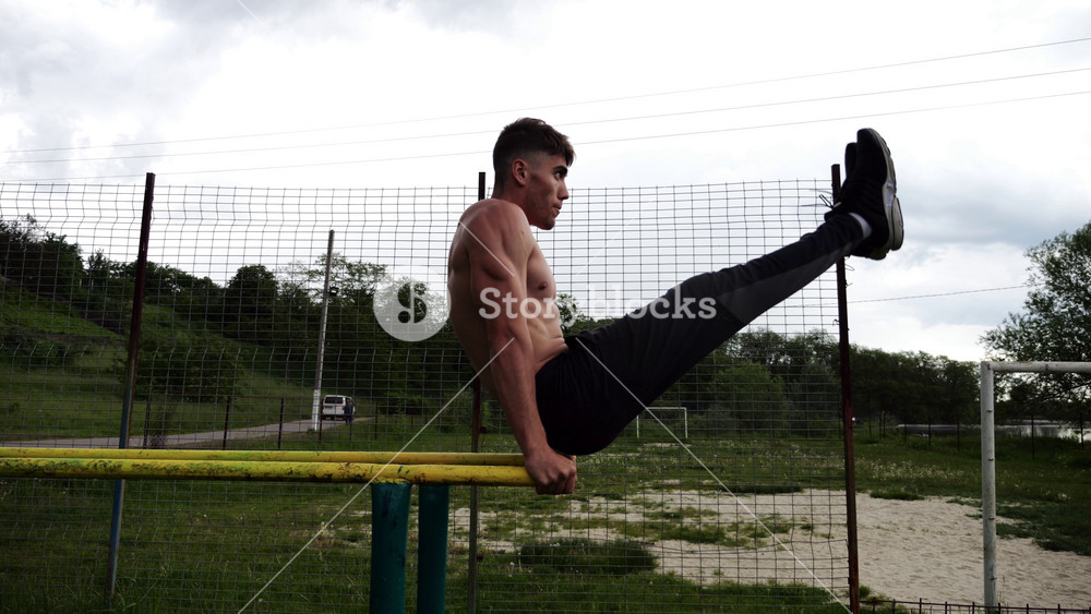 Strong man trains abdominal muscles raising legs on parallel bars at sports ground. Young shirtless sportsman exercising at nature. Sporty athlete working out at nature. Concept of sportive lifestyle