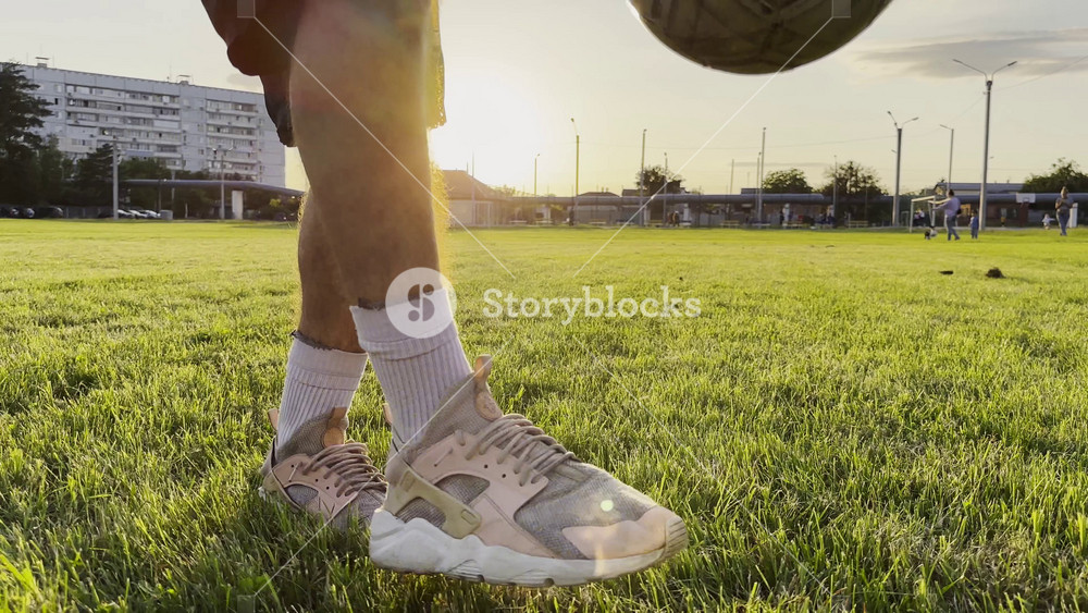 Male feet of professional footballer kicking ball at green field. Legs of young man juggling soccer ball on stadium at sunset. Sportsman