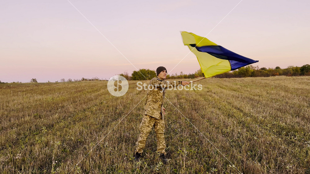 Male military lifted flag of Ukraine in honor of victory against russian aggression at dusk. Young soldier of ukrainian army waving blue