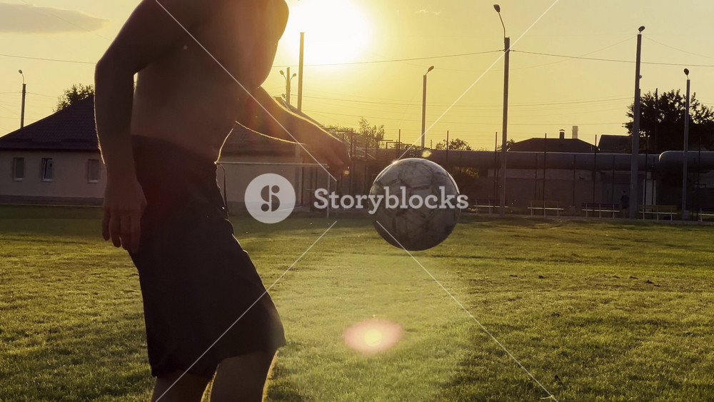 Young man juggling soccer ball on stadium at sunset. Professional footballer kicking ball at green field. Sportsman practicing tricks at